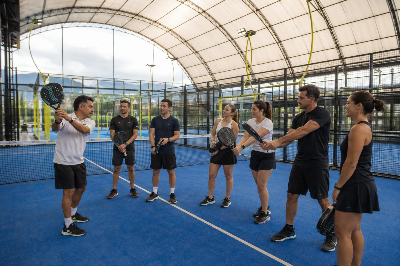 Instructor con raqueta explicando una jugada a un grupo de seis personas en una cancha azul cubierta
